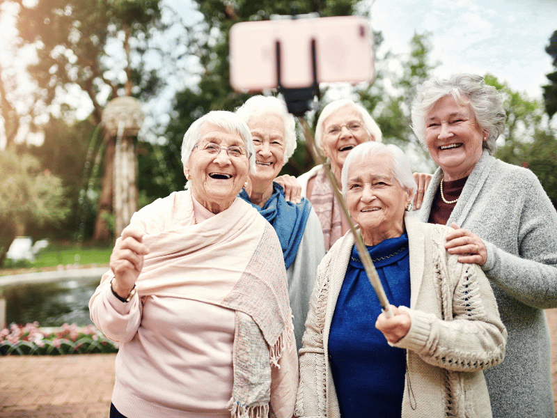 Dom seniora pięć starszych kobiet stoi blisko siebie na zewnątrz, uśmiechając się i pozując do selfie za pomocą smartfona na kijku do selfie, z zielenią i fontanną w tle. czarny dunajec