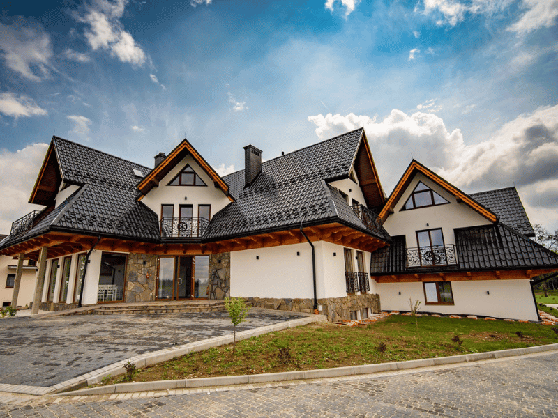 Dom seniora duży, nowoczesny dom ze stromymi dachami pokrytymi czarną dachówką, białymi ścianami i kamiennymi akcentami. dom ma kilka szczytów i balkonów i znajduje się na zagospodarowanym podwórku z wybrukowanym podjazdem pod częściowo zachmurzonym niebem. czarny dunajec