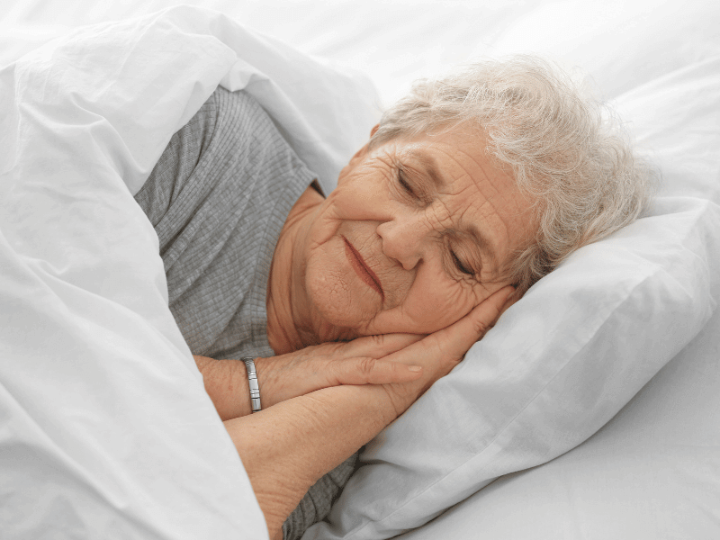 Dom seniora starsza kobieta z krótkimi siwymi włosami spokojnie śpi w łóżku, opierając głowę na białej poduszce i leżąc pod białym kocem. jej dłonie są delikatnie ułożone pod jej policzkiem. czarny dunajec