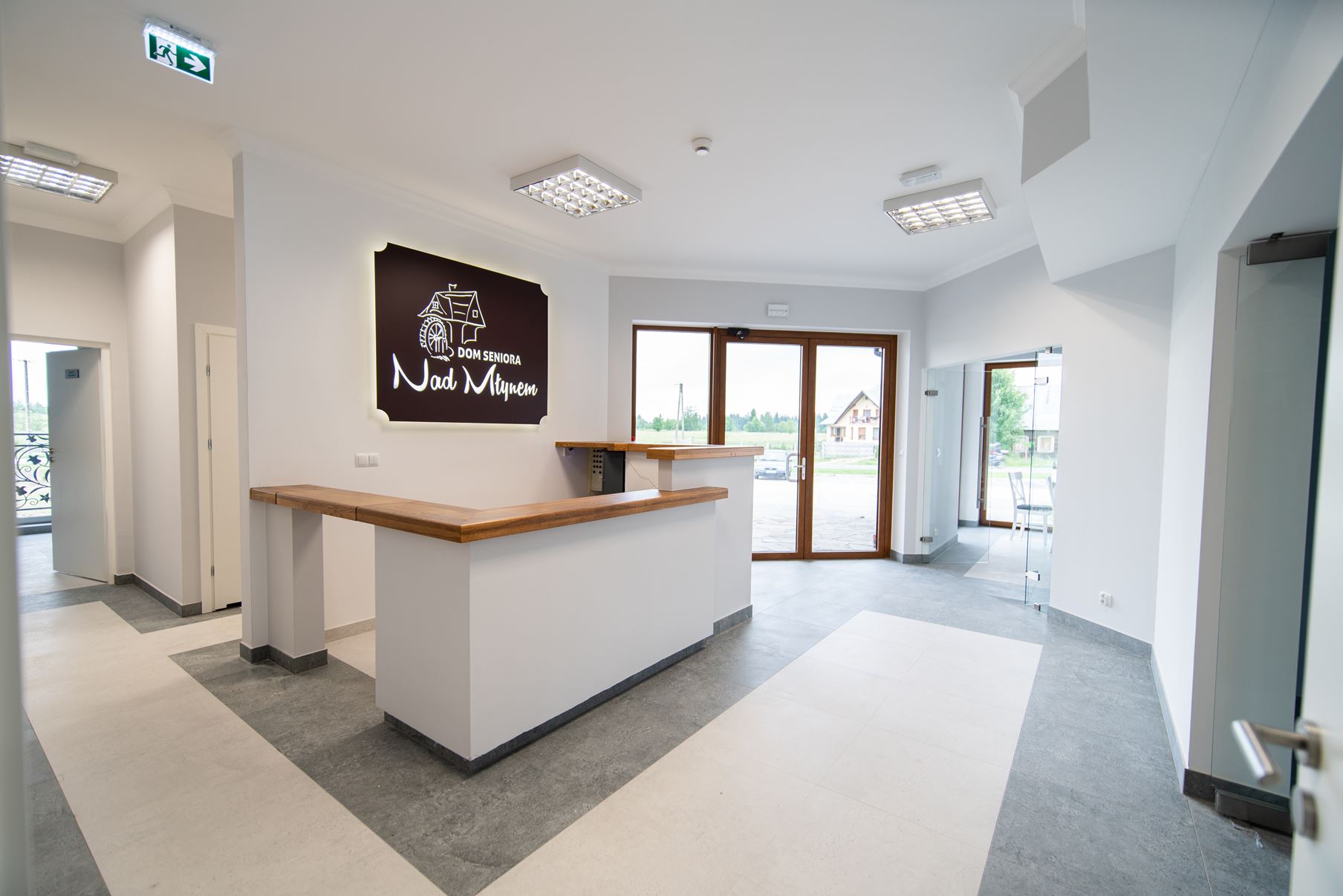 Dom seniora nowoczesna, jasna recepcja z drewnianą ladą, białymi ścianami, podłogą wyłożoną szarymi kafelkami, dużymi oknami i napisem na ścianie "nad młynem". drzwi i korytarze prowadzą do innych pomieszczeń. czarny dunajec